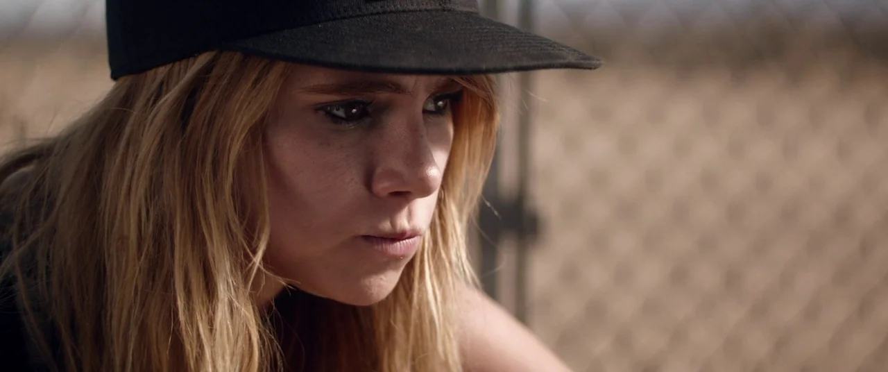 The Bad Batch (2016) movie still: closeup — A close-up shot of a young blonde woman standing near a chain link fence. She wears a black cap and…