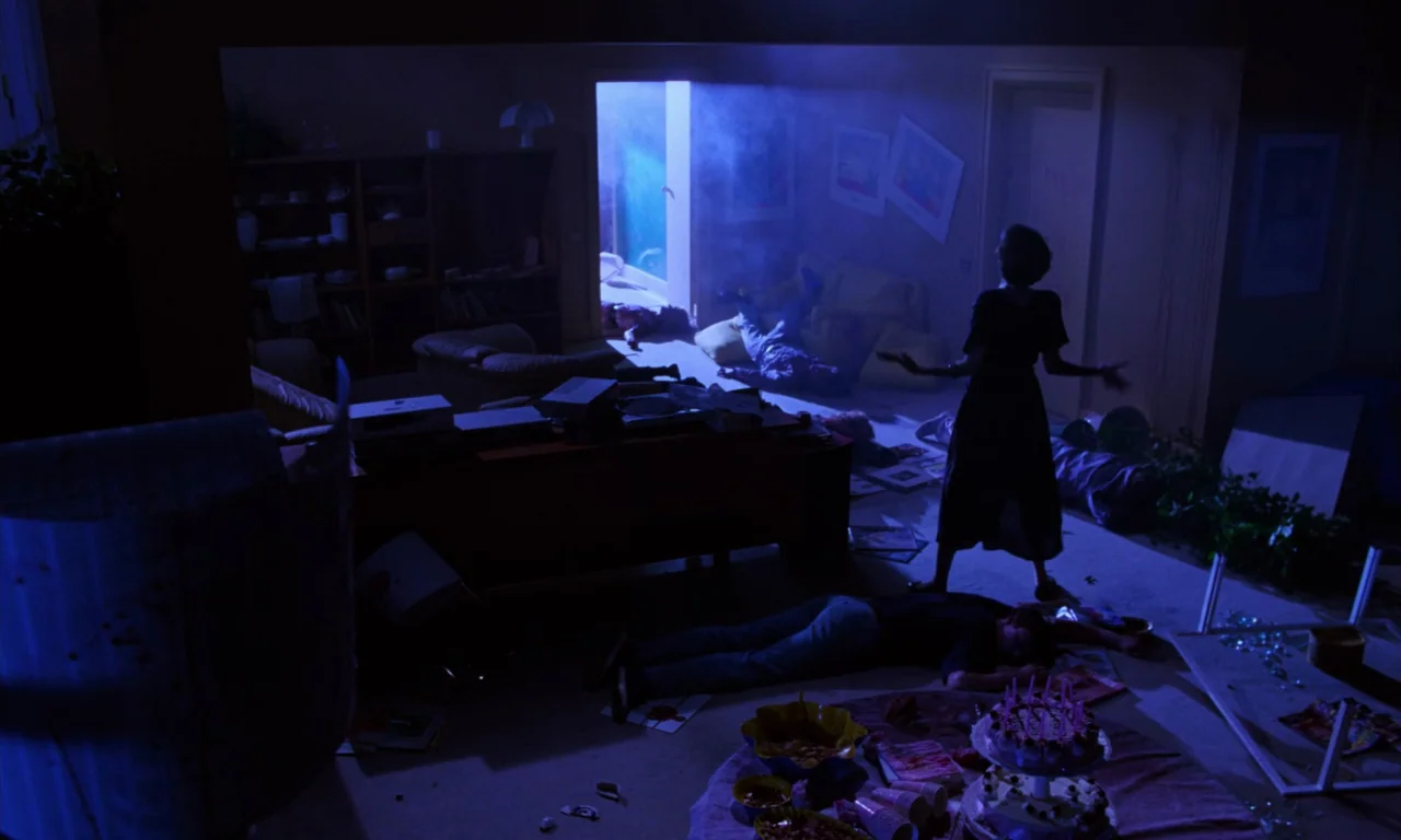Demons 2 (1986) movie still: wide — A cluttered room features multiple bodies lying on the carpet near a party table. A single woman stands near the…