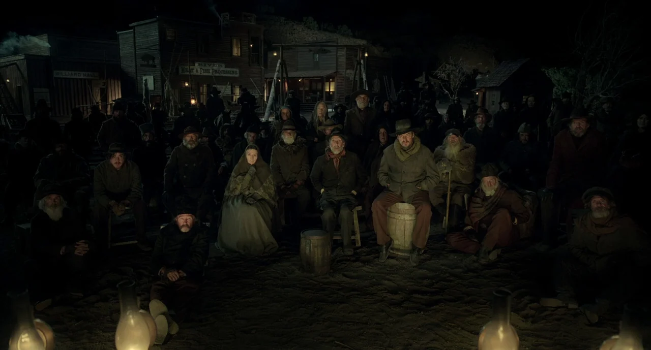 The Ballad of Buster Scruggs (2018) movie still: wide — A crowd of people in 19th-century attire sits in a dusty, dark town plaza. Wooden…