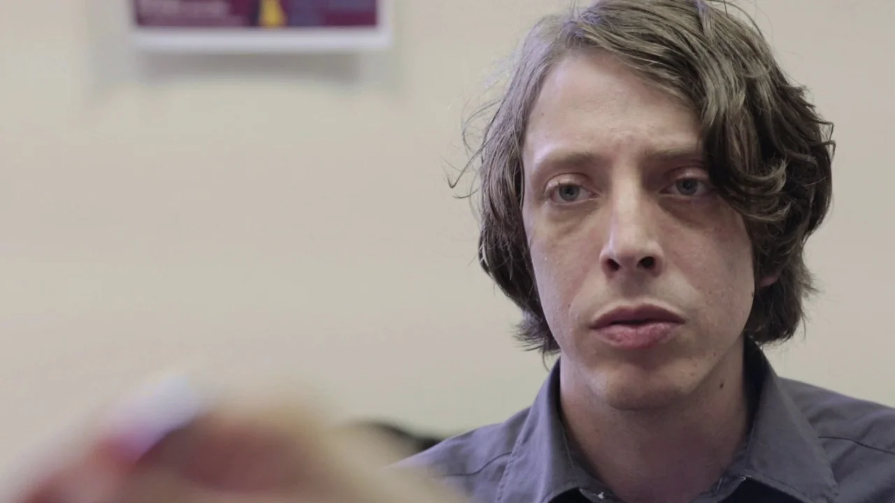 Buzzard (2014) movie still: closeup — A man sits in an office space with a pale wall. He looks towards the side while a blurred hand remains in the…