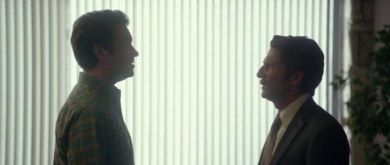 Faults (2014) movie still: medium — An interior space with bright vertical blinds covering a large window behind two men. The men stand in profile…