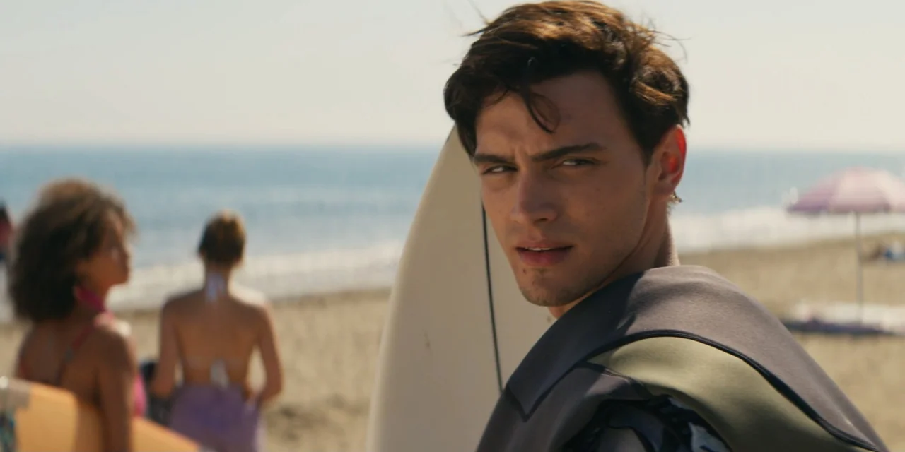 My Fault (2023) movie still: medium — A sunny beach with light sand and soft waves in the background. A young man with a surfboard stands in the…