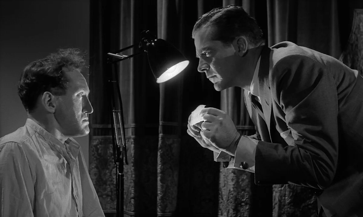 Night Of The Demon (1957) movie still: medium — A dark space features a desk lamp illuminating two men standing near curtains. One man holds a small…