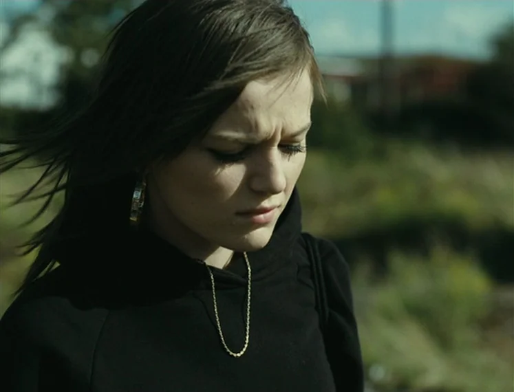 Fish Tank (2009) movie still: closeup — An outdoor field setting with blurred vegetation visible in the background under daylight. A young woman…