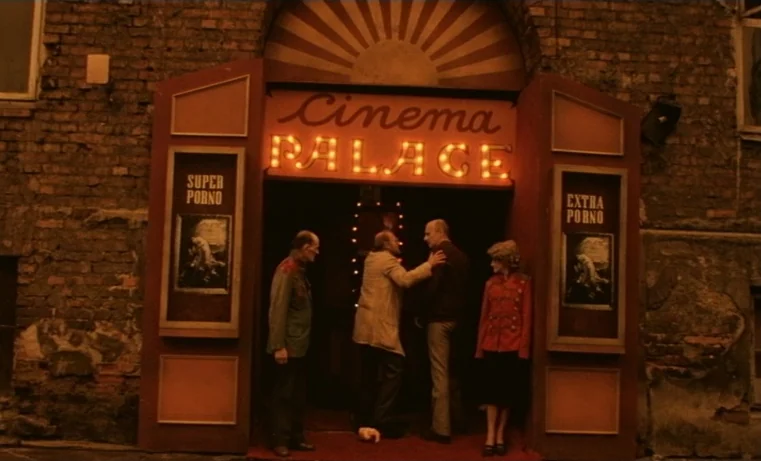 Golem (1980) movie still: fullbody — A brick-walled storefront entrance displays a neon sign reading Cinema Palace. Four people stand before the…