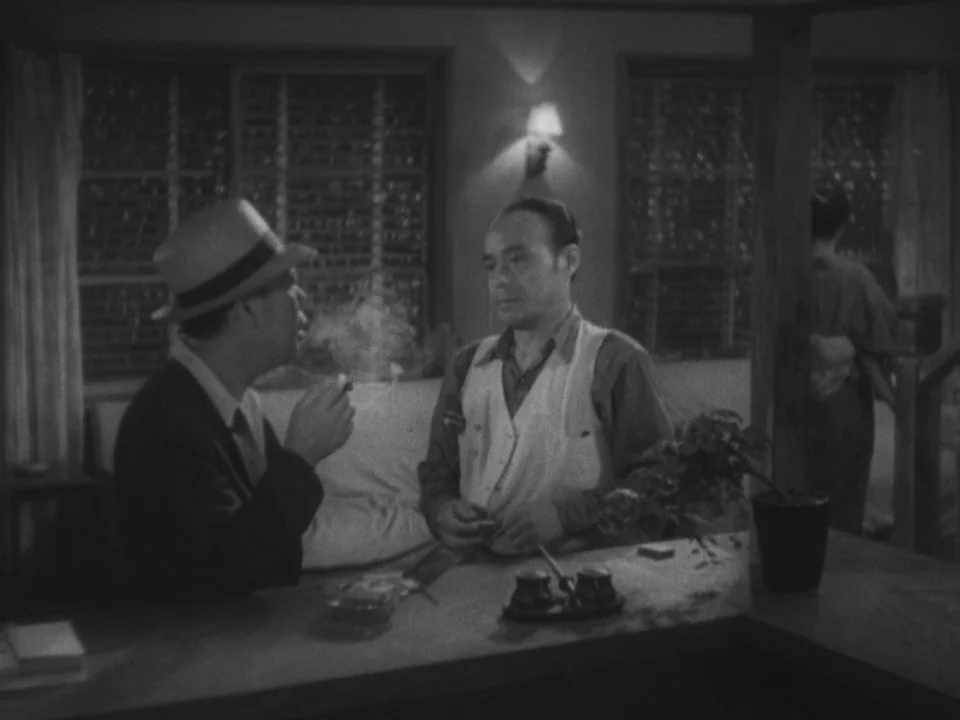 Stray Dog (1949) movie still: medium — Two men sit at a counter inside a dimly lit space with wood paneled walls. The man on the left wears a hat…