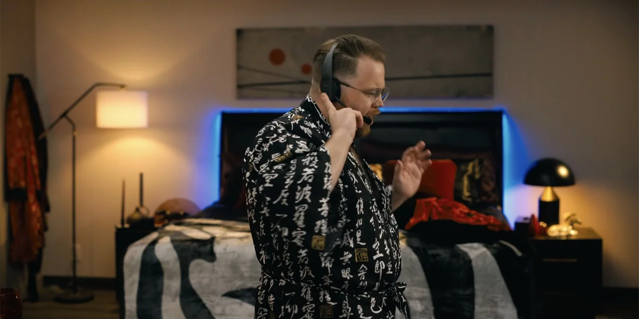 Playdate (2025) movie still: medium — A modern bedroom contains a dark bed frame with blue LED lighting strips behind it. A man wearing a black and…