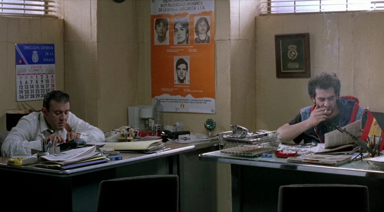 Kika (1993) movie still: wide — A dimly lit room functions as a workspace with beige walls and cluttered desks. Two men sit across from each other,…