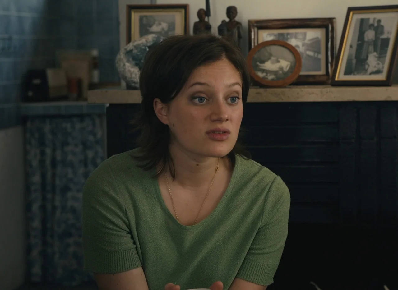 Red Island (2023) movie still: medium — A residential room features a fireplace mantle decorated with multiple framed photographs. A young woman in…