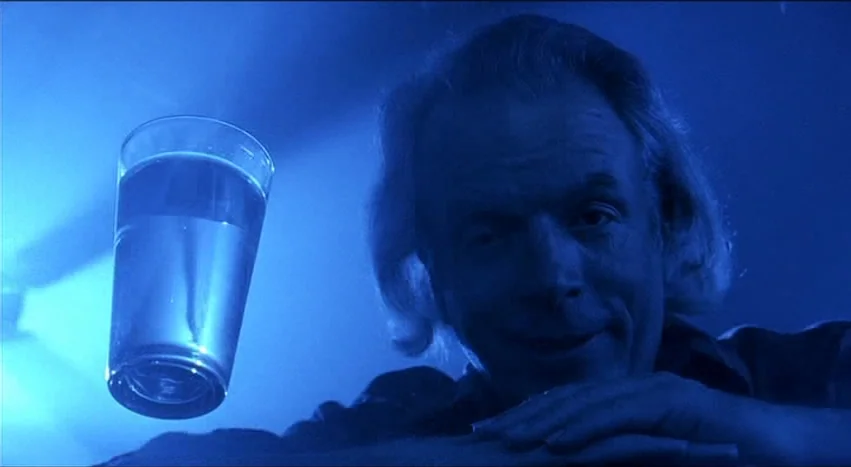 Gray’s Anatomy (1996) movie still: closeup — An older man looks toward the camera while resting his arms on a surface. A glass filled with water is…