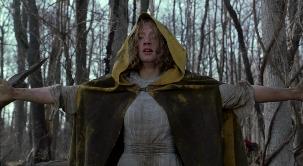 The Village (2004) movie still: medium — A dense forest of leafless trees provides a stark backdrop for a woman in a cloak. A woman stands in the…