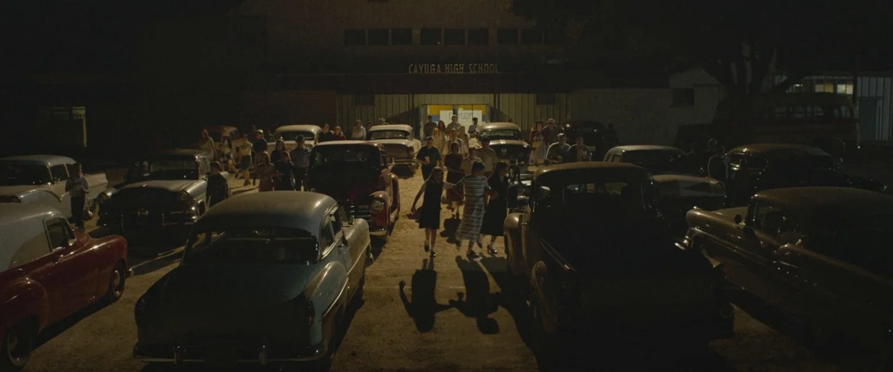 The Vast of Night (2019) movie still: wide — A dark, dirt-covered parking lot is filled with rows of classic mid-century automobiles. Several…