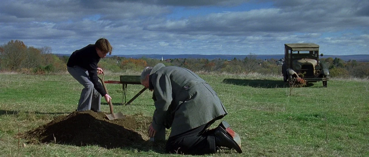The Cider House Rules movie still: wide — A vast, open field with rolling hills and a distant town under a cloudy sky. An elderly man kneels beside…
