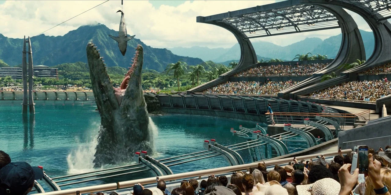 Jurassic World (2015) movie still: wide — A modern outdoor aquatic amphitheater features tiered concrete seating packed with onlookers under a…