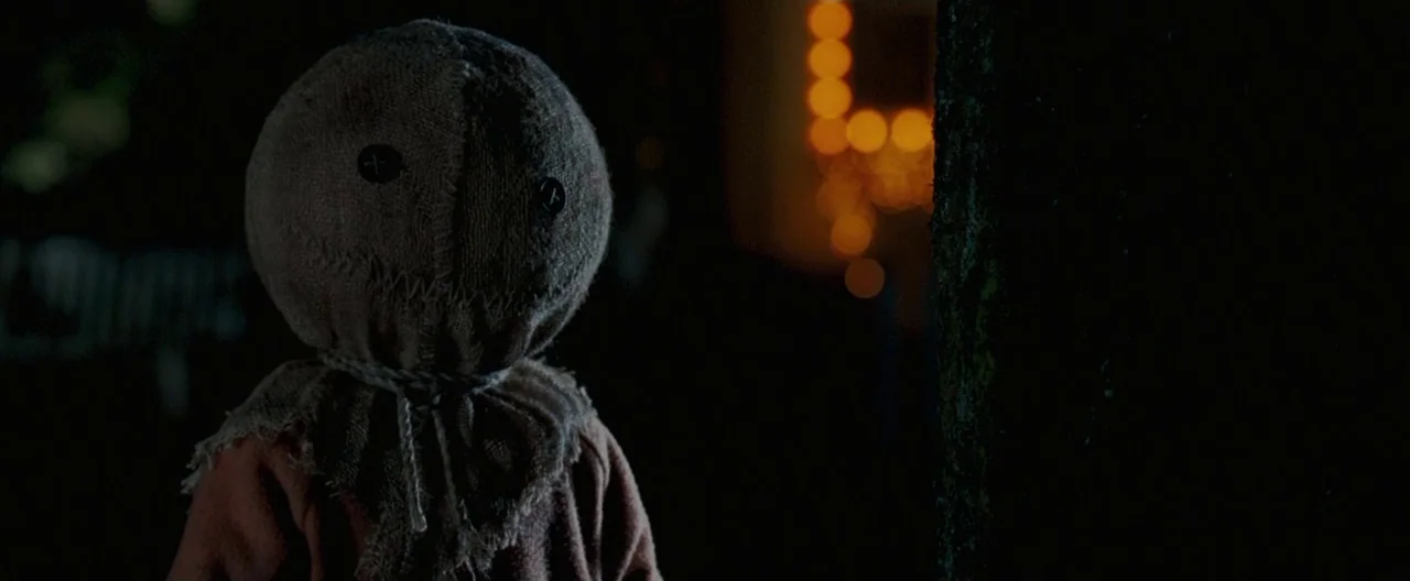 Trick ‘R Treat (2007) movie still: closeup — A nighttime outdoor setting with a dark tree trunk on the right side. A small figure wearing a burlap…