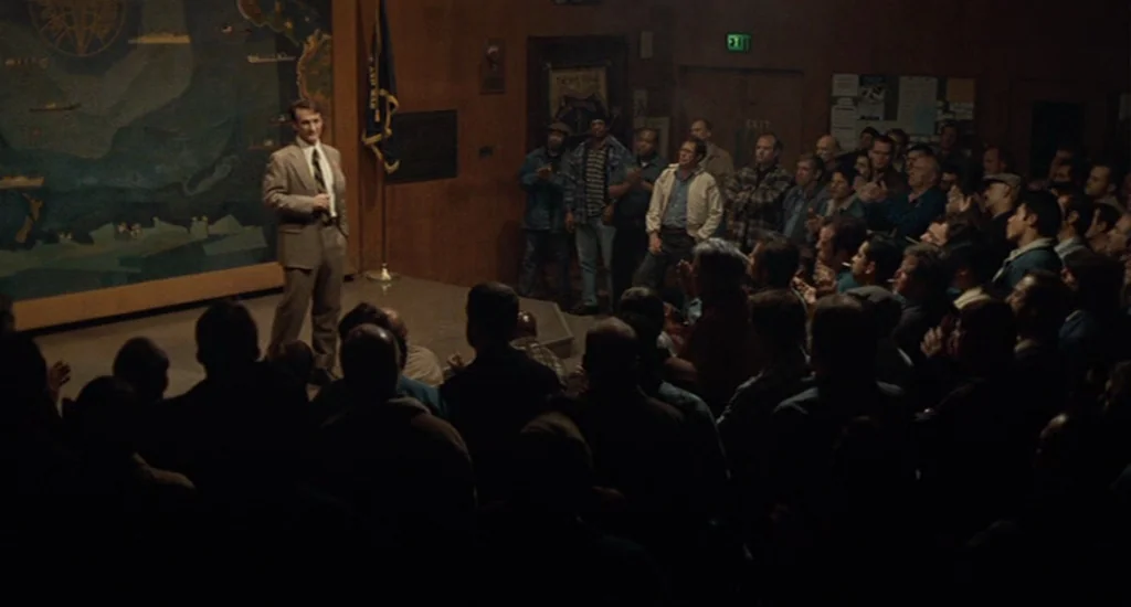 Milk (2008) movie still: wide — A dimly lit union hall with wood-paneled walls features a large map on display. A man stands on a raised platform…