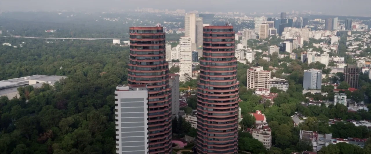 Do It Like An Hombre (2017) movie still: establishing — An aerial view shows two prominent cylindrical red towers rising above a dense urban forest.…
