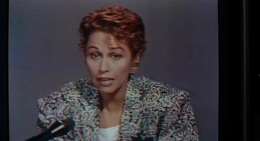 Matador (1986) movie still: medium — A woman sits in a television studio set against a featureless grey backdrop. She is looking directly at the…