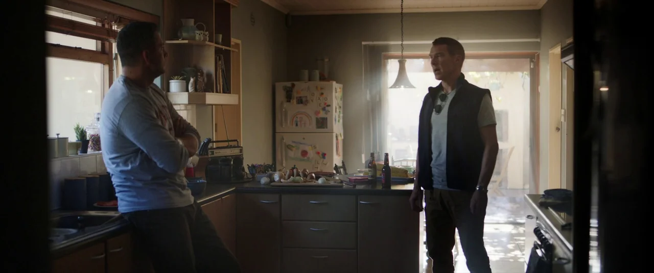 The Mauritanian (2021) movie still: fullbody — A residential room contains a refrigerator covered in magnets and a central island workspace. Two men…
