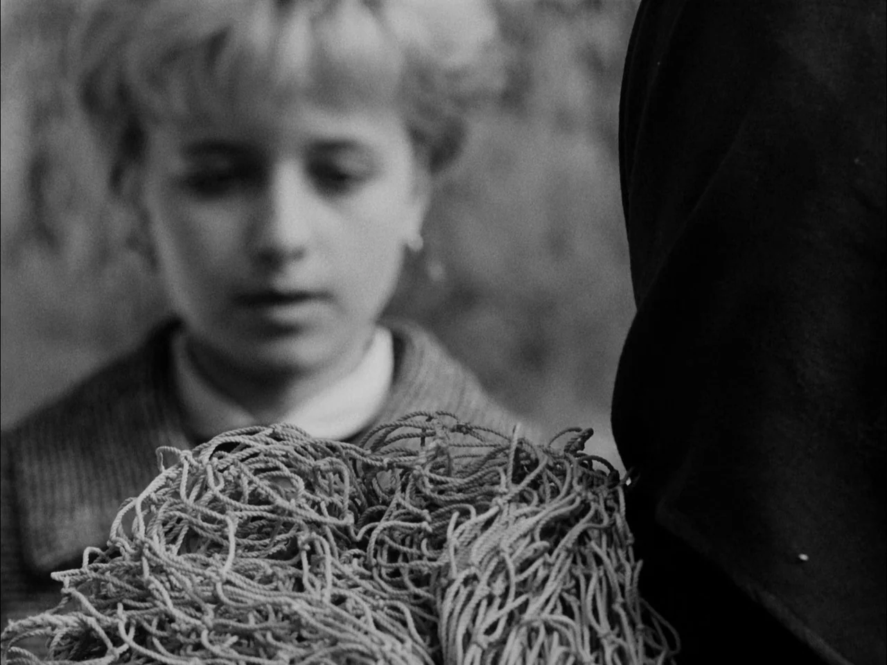 The Sun In A Net (1963) movie still: medium — A person holds a large, tangled mass of fishing netting in the foreground. A child standing nearby…