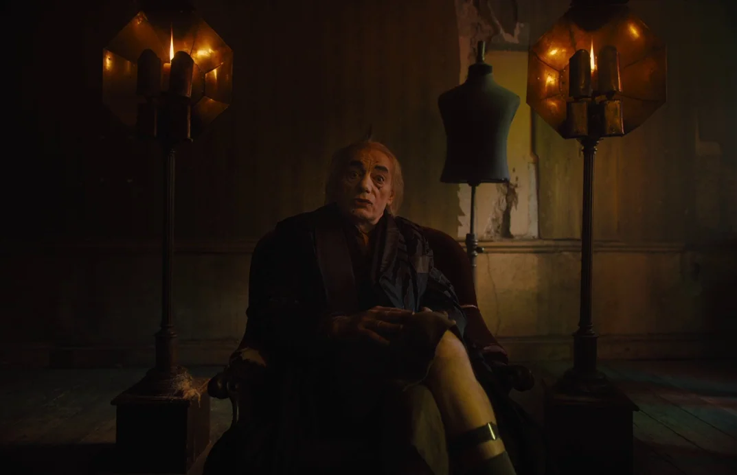 Gretel & Hansel (2020) movie still: medium — A dimly lit room contains two ornate metallic floor lamps and a dressmaker's dummy. An elderly man sits…