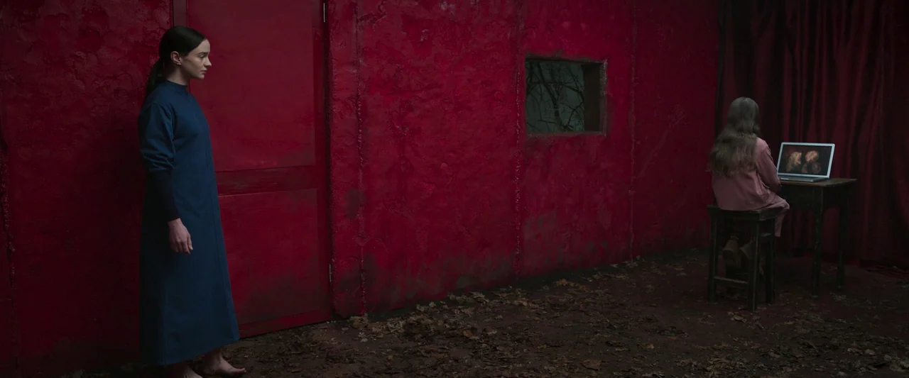 Stopmotion (2024) movie still: wide — A stark room features a rough, textured deep red wall with a small recessed window. A woman stands on the…