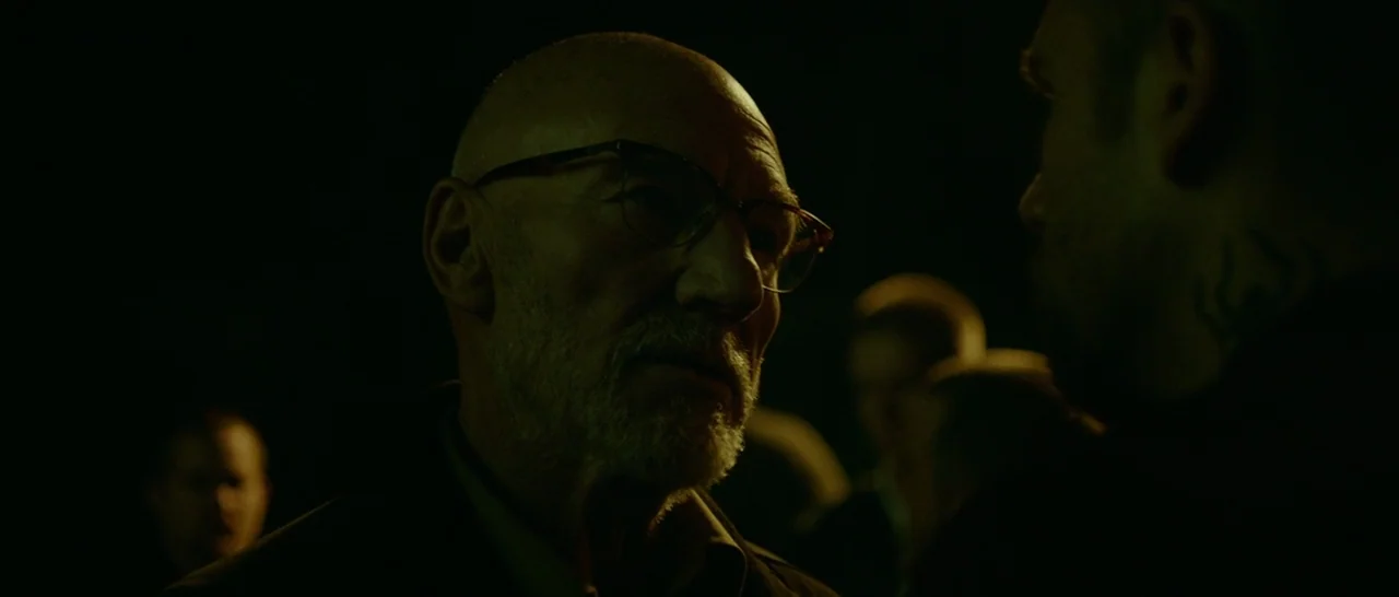 Green Room (2015) movie still: closeup — A dark outdoor environment is lit by a dim, greenish light source at night. An older man with glasses…