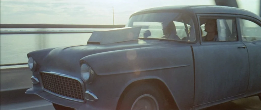 Two Lane Blacktop (1971) movie still: medium — A matte grey 1955 Chevrolet with a hood scoop drives across a large bridge. Two young men with long…
