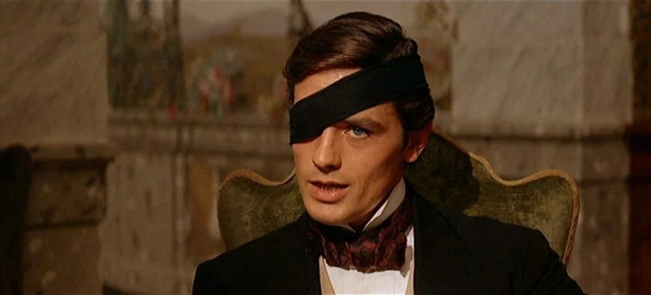 The Leopard (1963) movie still: closeup — A refined interior with a faint wall mural visible behind a dark chair. A young man wearing a black eye…