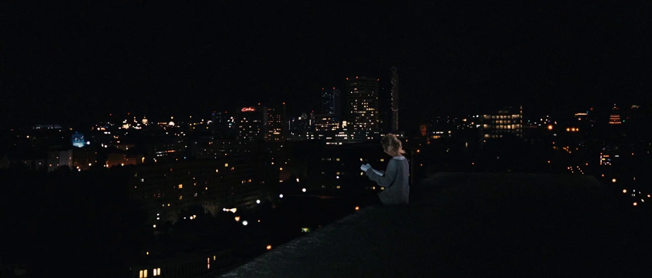 Sound of Noise (2010) movie still: wide — A dark city skyline viewed at night from a high vantage point. A person sits on the roof edge reading a…
