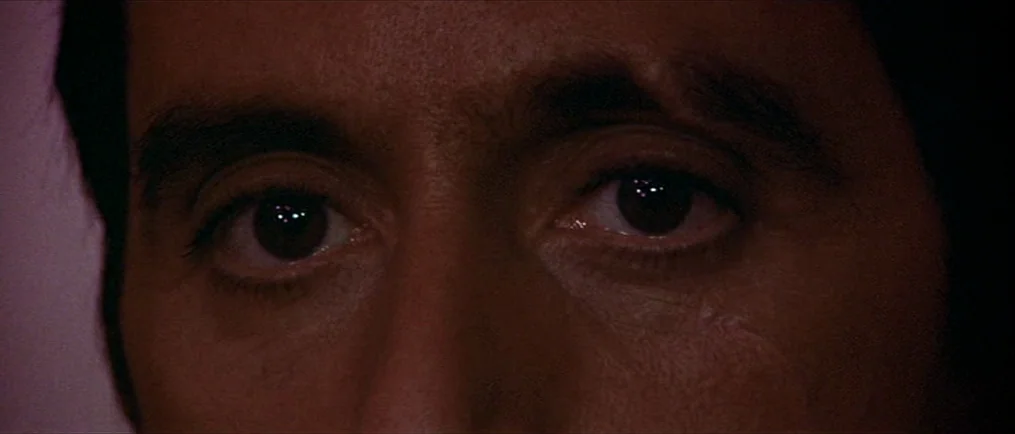 Scarface (1983) movie still: closeup — An extreme close-up focuses on a man's eyes and brow. The face is shrouded in deep shadow around the edges of…