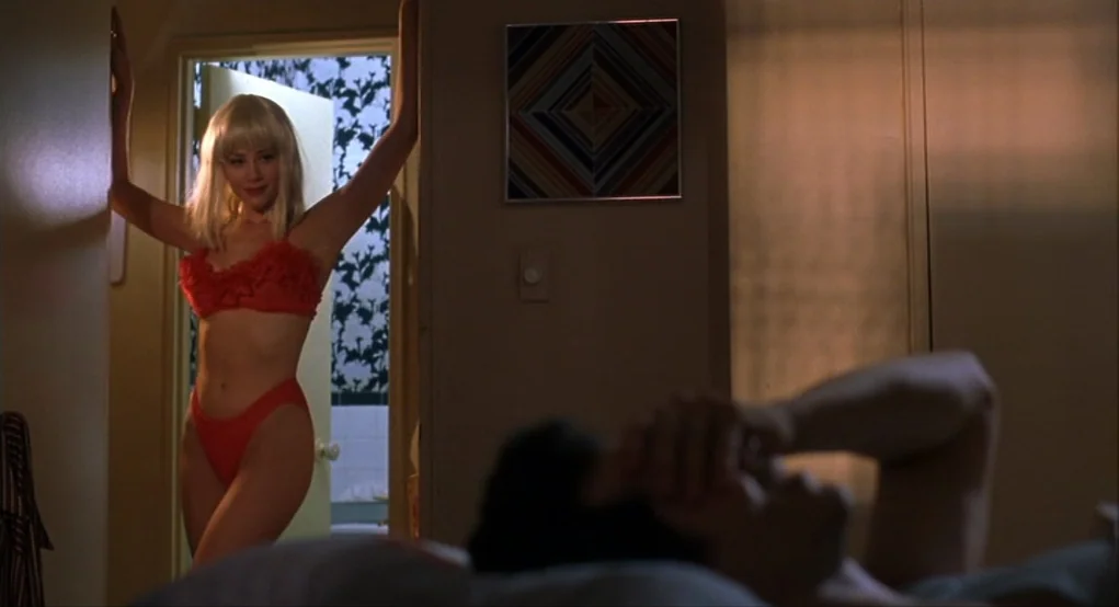 Summer of Sam (1999) movie still: medium — A dimly lit room with brown walls and a small colorful geometric wall art. A woman in a red bikini stands…