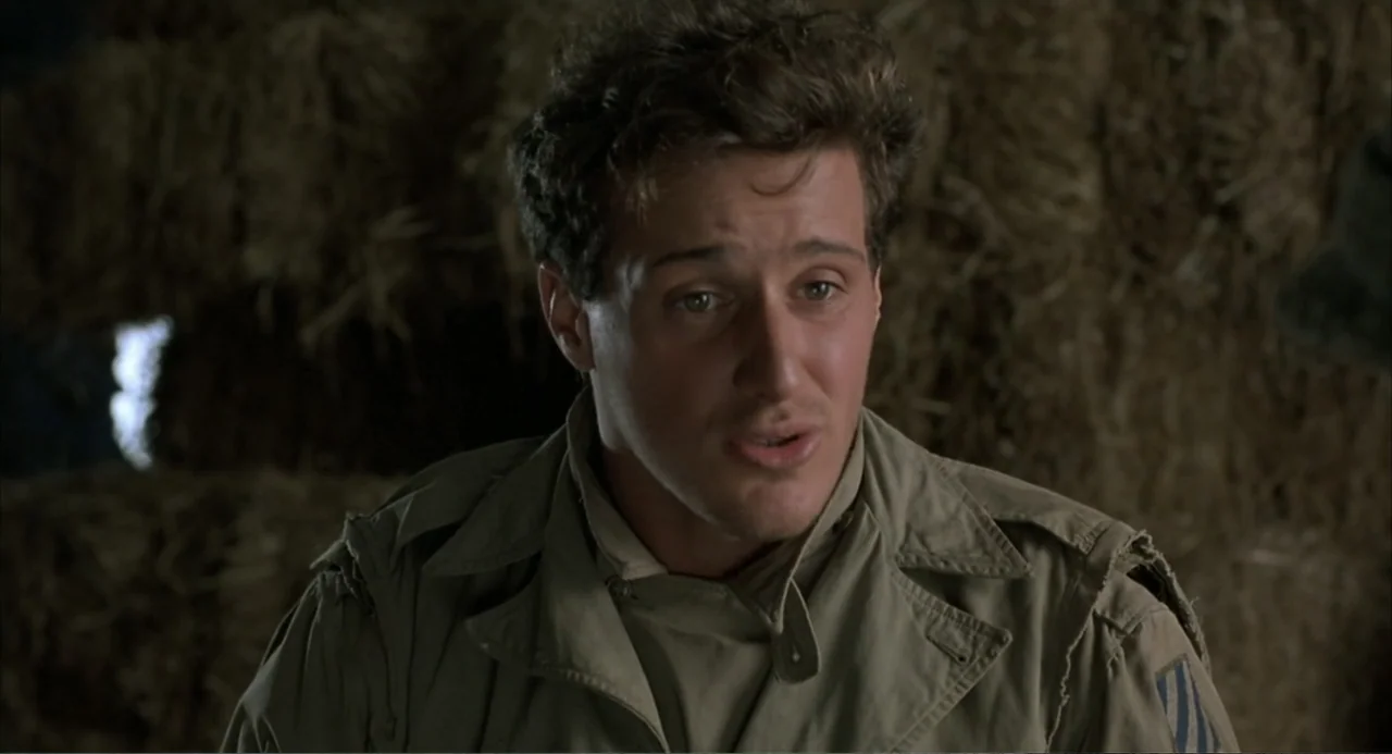 Zone Troopers (1985) movie still: medium — The interior of a rustic barn features stacks of hay as a backdrop. A young soldier in a military uniform…