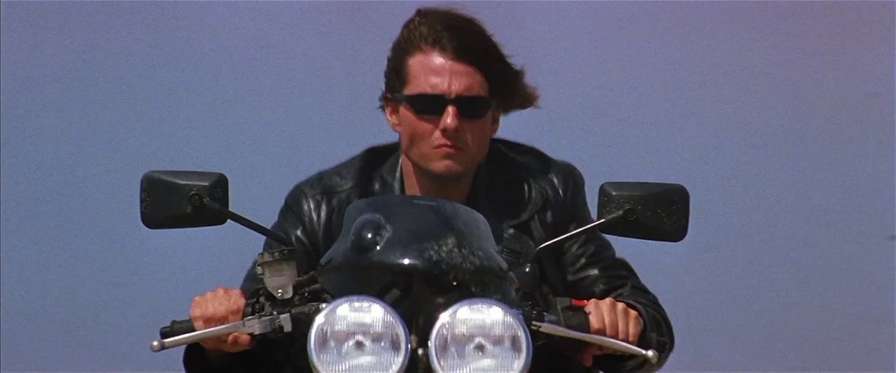 Mission: Impossible II (2000) movie still: medium — A man rides a motorcycle against a clear, expansive sky backdrop. He holds the handlebars while…