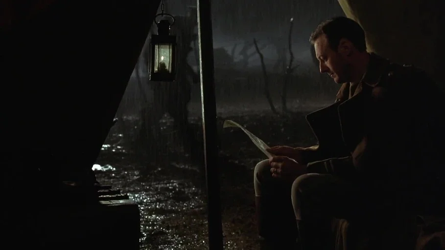 A Little Princess (1995) movie still: medium — A man sits inside a dark canvas military tent during a rainstorm at night. He is reading a letter by…