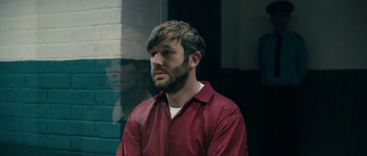 Calvary (2014) movie still: medium — A man in a red uniform sits in front of a glass partition. A uniformed guard stands in the shadows in the…