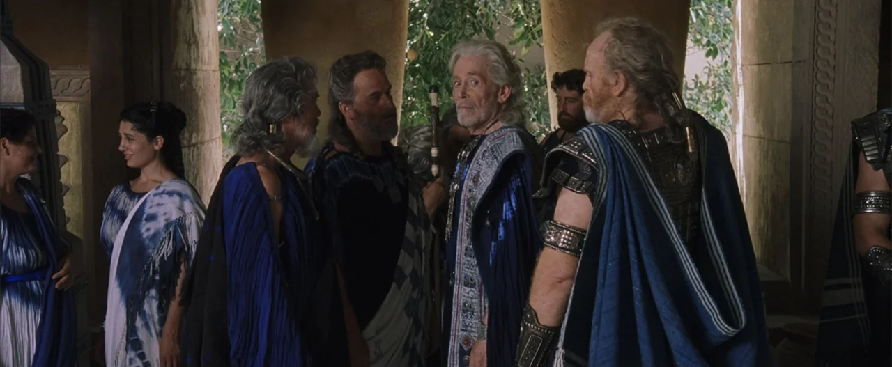 Troy (2004) movie still: fullbody — A group of people in traditional ancient robes stand beneath a stone colonnade outdoors. Several men and women…