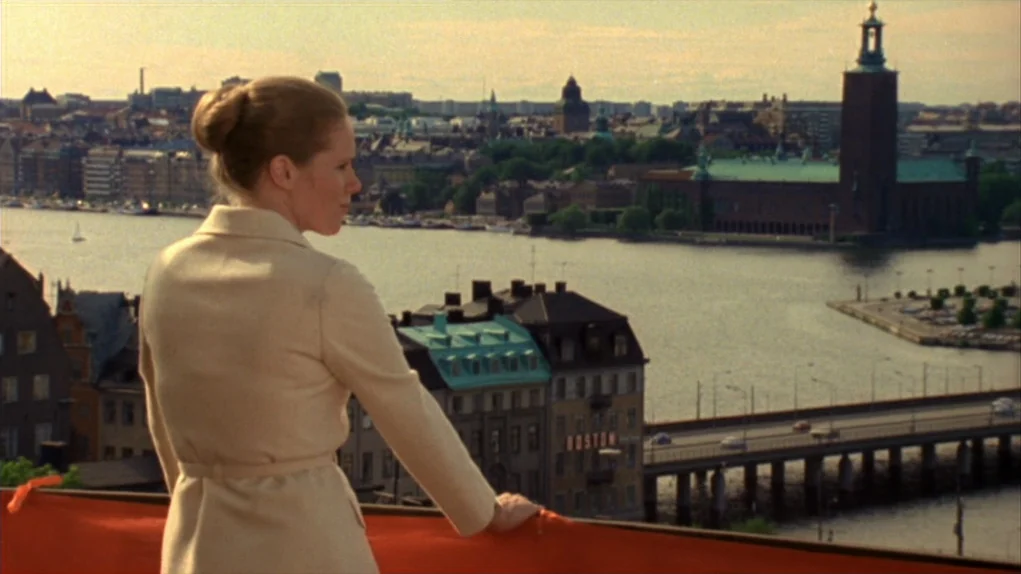 Face to Face (1976) movie still: medium — A woman stands on a balcony overlooking a city landscape with a prominent waterfront. The city includes a…