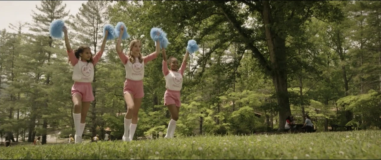 Summer Camp (2024) movie still: fullbody — A sunny grassy park is populated by tall mature trees and a picnic area. Three teenage cheerleaders in…