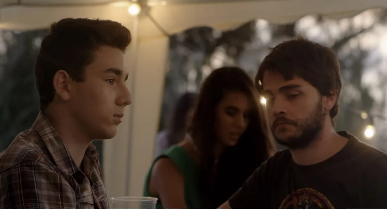 Newton's Third Law (2017) movie still: medium — An outdoor gathering under a tent with hanging string lights at dusk. Three young adults are sitting…
