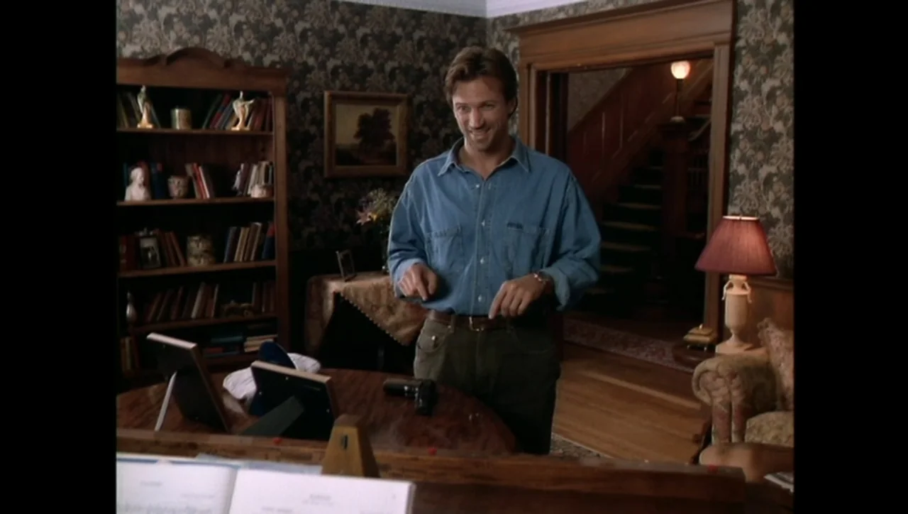 The Awakening (1995) movie still: medium — A residential living room features wood paneling, ornate wallpaper, and a staircase in back. A man smiles…