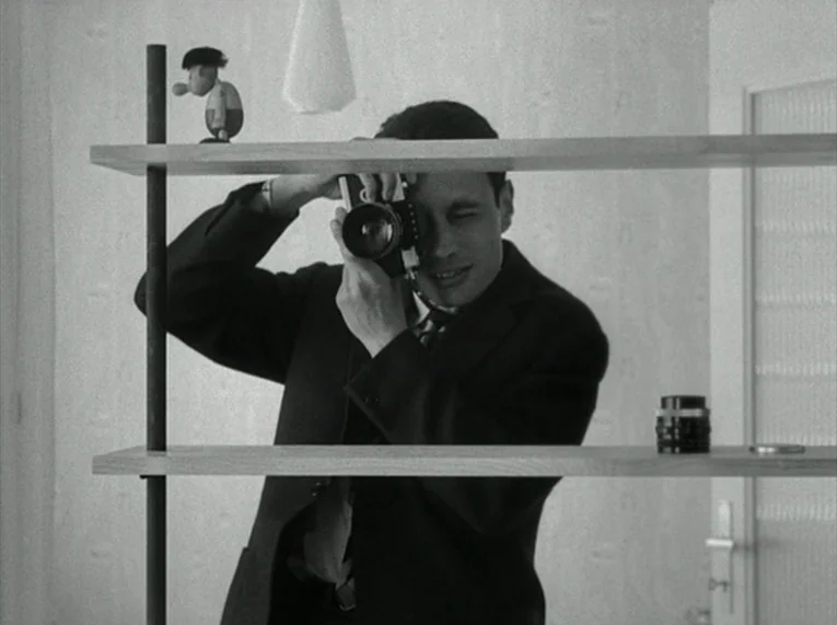 Le Petit Soldat (1963) movie still: medium — A man stands behind a wooden shelving unit in a bright minimalist apartment. He holds a 35mm camera up…