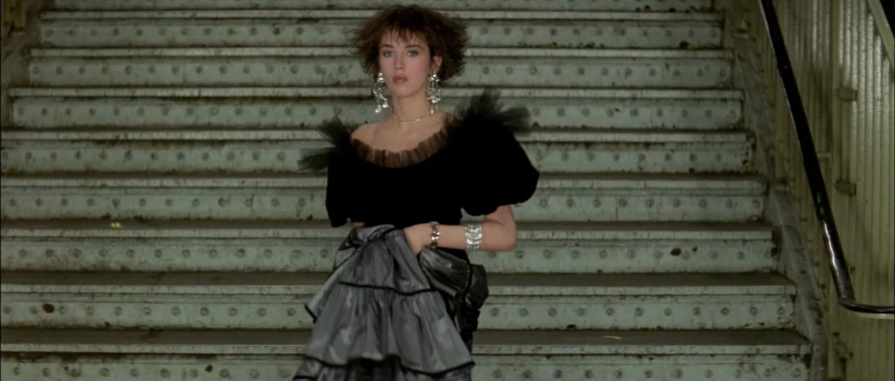Subway (1985) movie still: medium — A set of industrial concrete steps with metal trim background a young woman. The woman stands facing forward,…