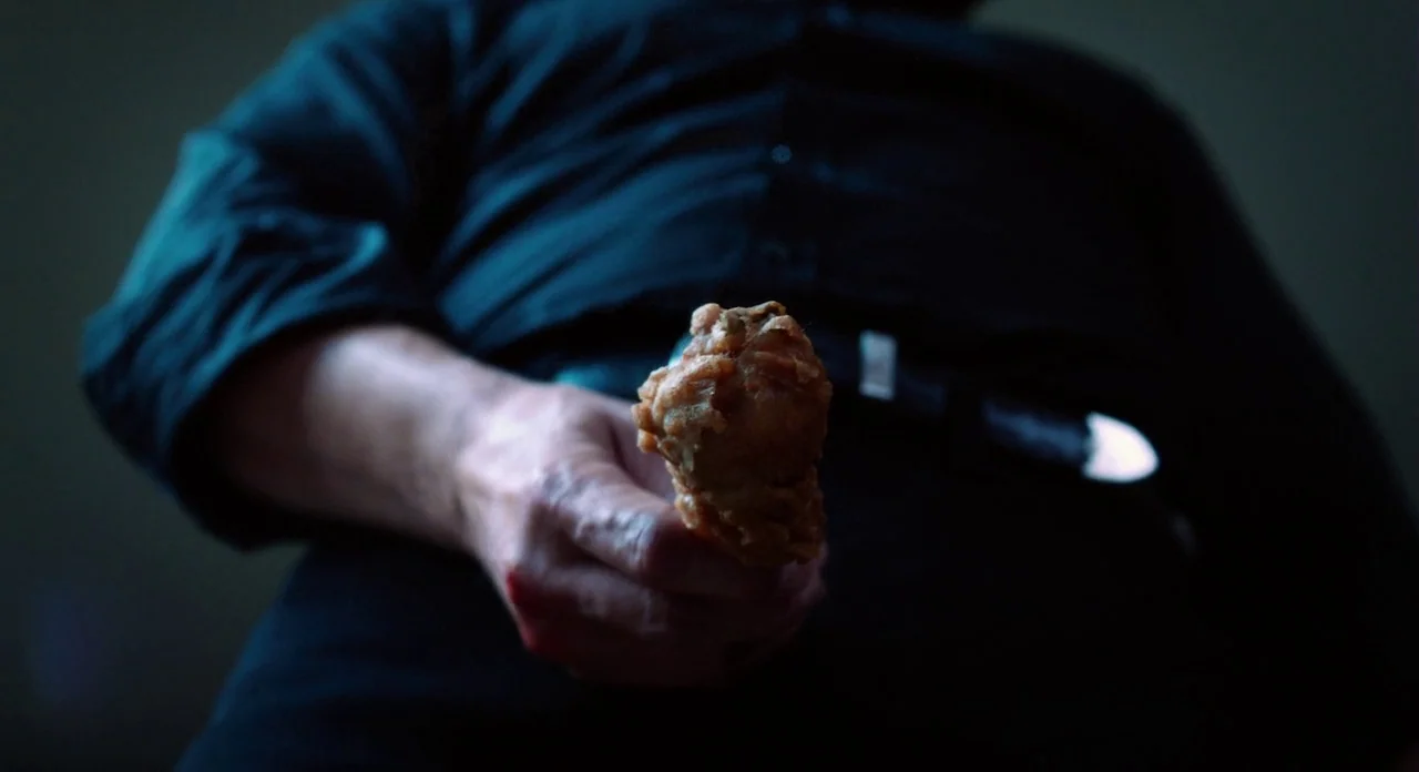 Killer Joe (2011) movie still: medium — A dimly lit interior space with a person dressed in dark clothing. The subject holds a single piece of fried…