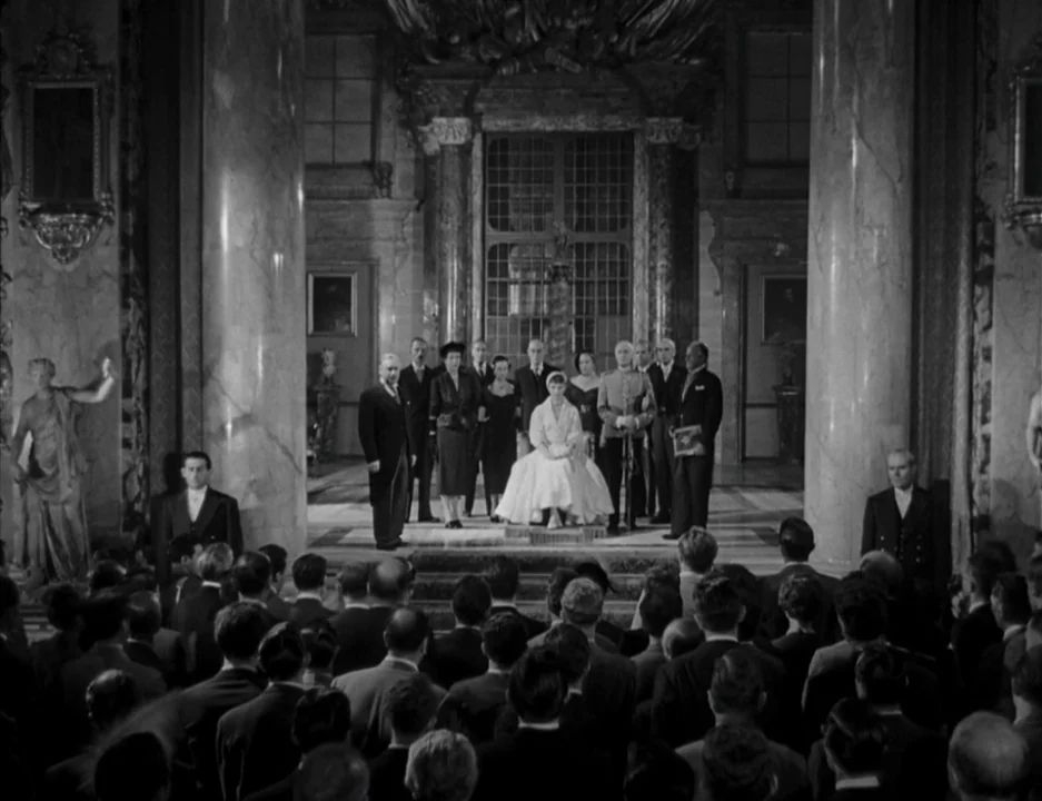 Roman Holiday (1953) movie still: wide — A grand stone-columned hall features an elevated dais with a seated woman in white. A large audience stands…