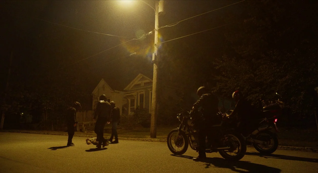 The Art of Self Defence (2019) movie still: wide — A dark residential street is illuminated by a single yellow streetlamp at night. Several figures…