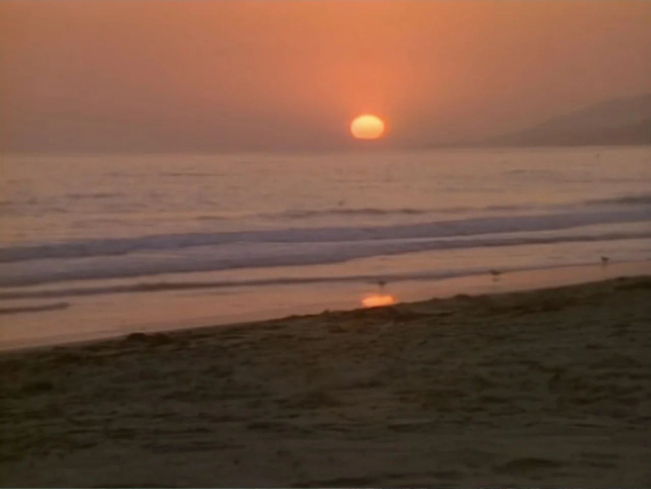 My First Love (1988) movie still: establishing — A quiet beach shoreline meets the ocean during a vibrant sunset with orange light. Small waves…