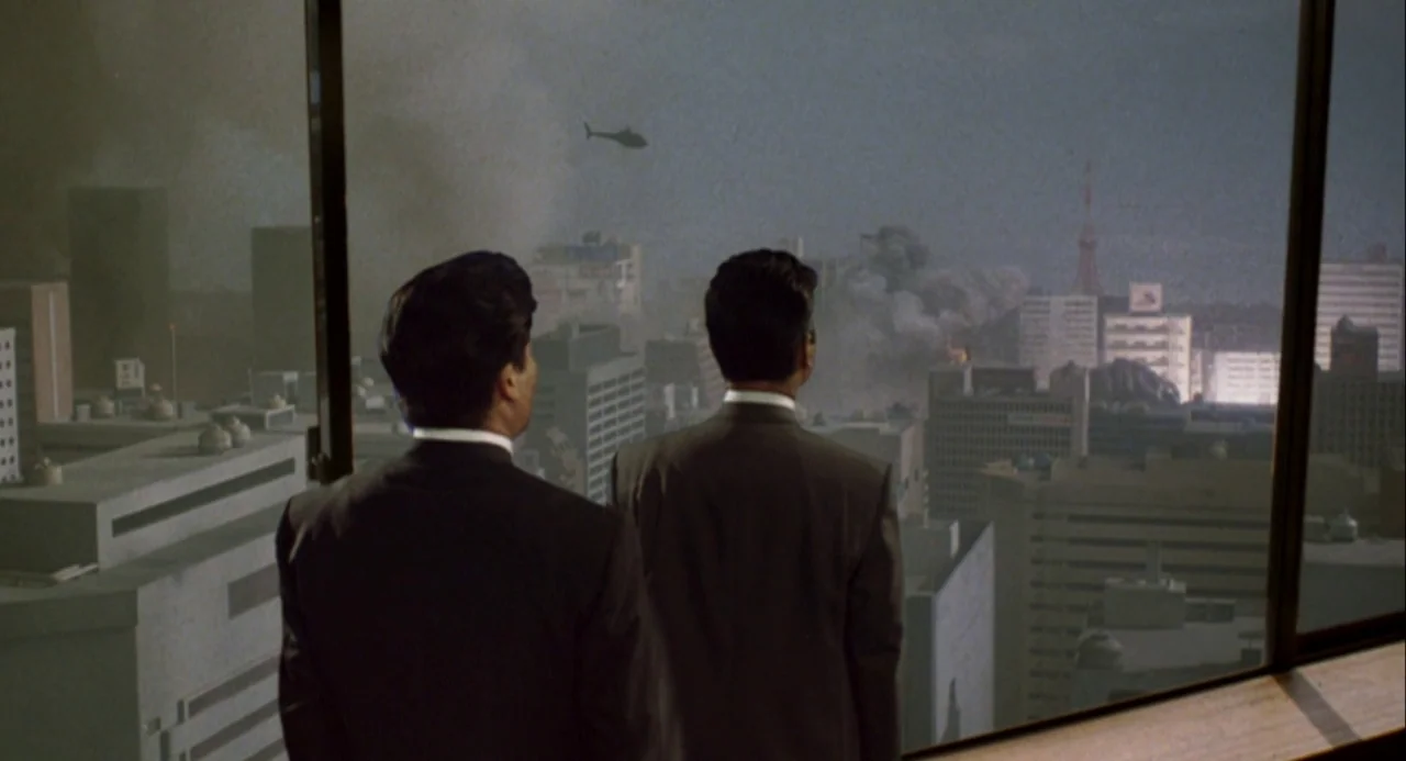 Godzilla and Mothra: The Battle for Earth (1992) movie still: medium — Two men in suits stand looking out a window at an urban landscape. Thick…