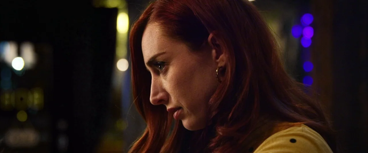 The Stylist (2020) movie still: closeup — A dark indoor space at night featuring blurred light bokeh in the background. A woman with long auburn…