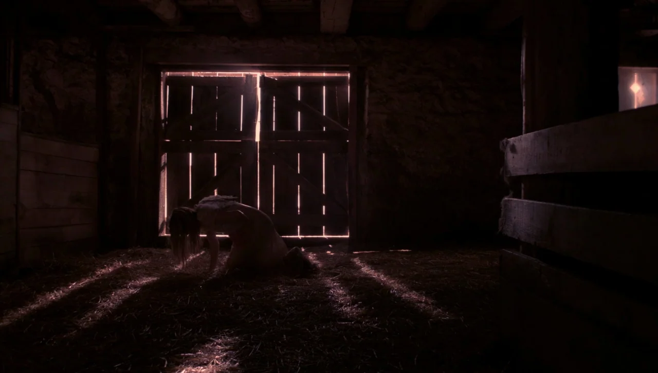 Hellions (2015) movie still: fullbody — A dark wooden barn interior has rough stone walls and a floor covered in straw. A woman kneels in the center…